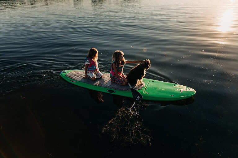 Two girls and a dog ride a stand up paddle board in the lake