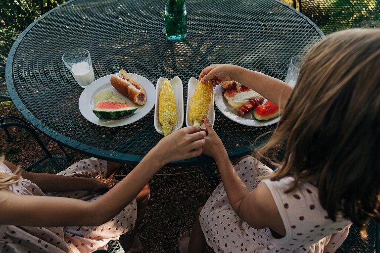 Two girls reach for a piece of corn on outdoor table. 