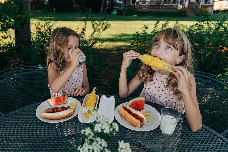 Girl drinks milk while her sister eats corn. 