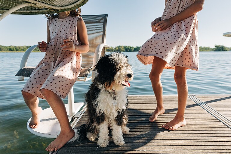 Dog sits on dock next to two girls in matching dresses. 