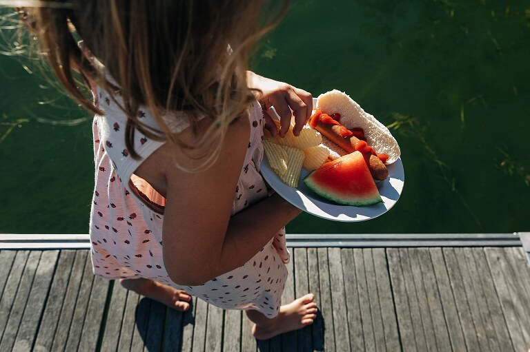 Girl eats chips, watermelon and a hotdog next to the lake. 
