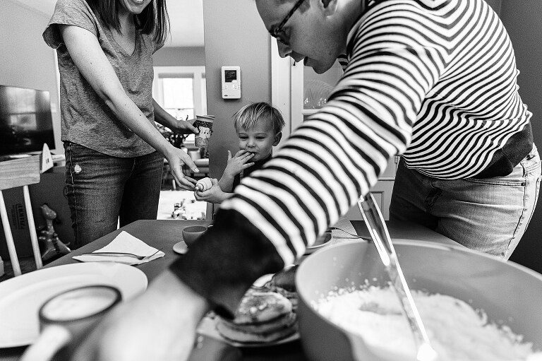 dad reaches for food while mom hands son food and boy puts fingers in his mouth on Saturday pancakes 