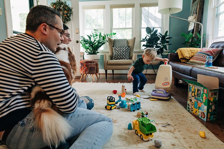 toddler boy uses play vacuum to clean up his toys while parents look on in amusement