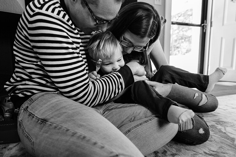 black and white. parents snuggle toddler son on their laps while sitting on the floor in their living room