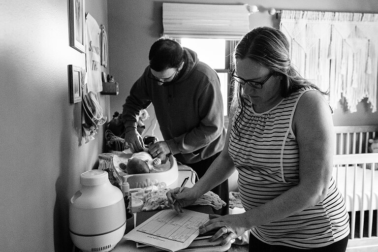 mom charts newborn needs while dad is in background changing baby's diaper