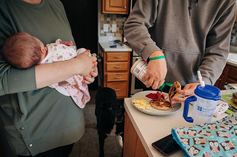 parents stand near each other while mom holds baby and dad pours syrup on mom's pancakes 