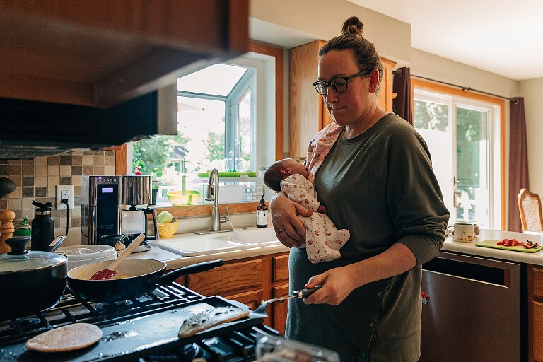 mom holds newborn while flipping pancakes with the other hand