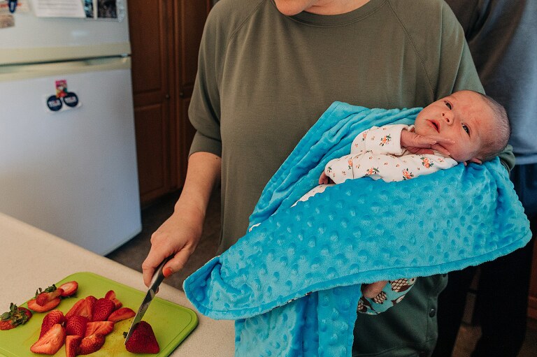 mom slices strawberries in non-dominant hand while holding newborn with her other hand