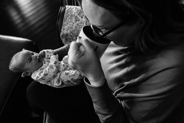 black and white. Mom sips coffee while newborn looks up from lap