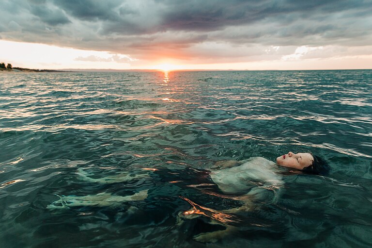 A preteen child lays still in shallow water, only face above water, while the sun sets behind him.