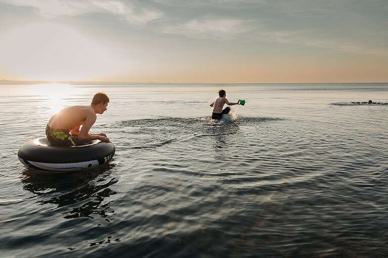Three boys play in the lake, forming a diagonal with their bodies as they swim toward the horizon line with the sun setting 