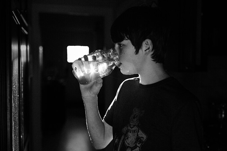 black and white. Boy drinks water from a Mason jar while backlit from the sun. His fingers and sunflare are visible through the drinking jar and his body is outlined with rim light 