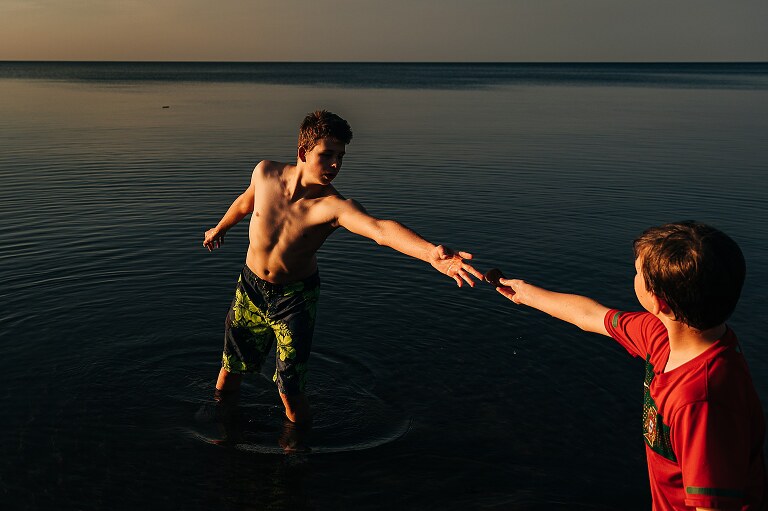 two boys standing in the lake form a diagonal with their arms as they hand off a skipping stone 