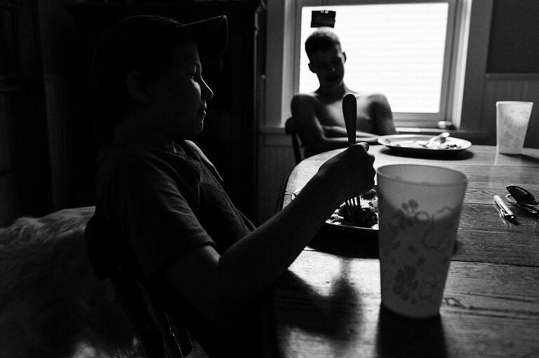 black and white. Boy two boys sit at dining room table. One boy is all in shadows with rim light on profile, one boy is silhouetted against bright window 