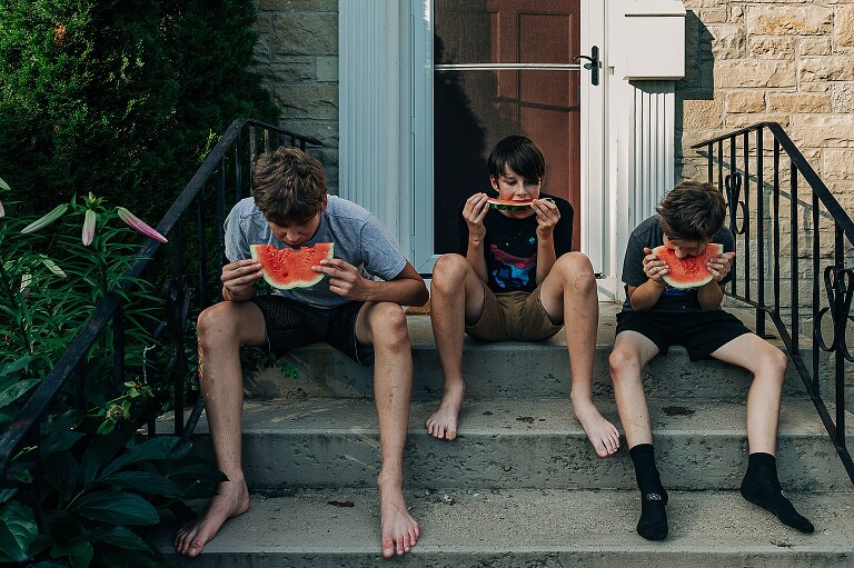 three teen boys sit on front stoop eating watermelon on summer's day