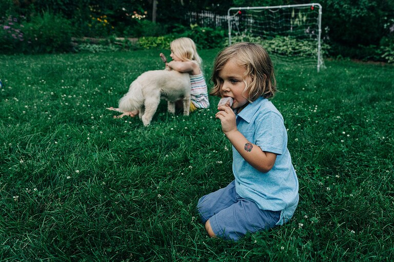 foreground: child eats FudgePop popsicle. Background: older child hugs small dog while holding FudgePop
