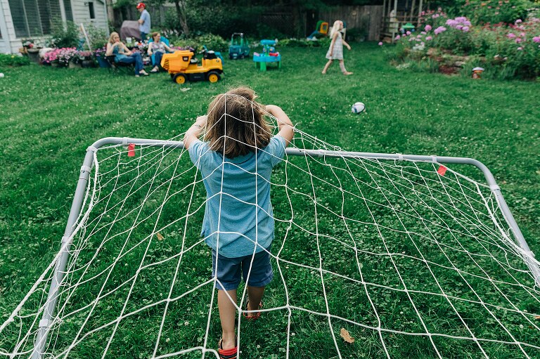 young child pulls a soccer net over his head while sibling, parents, and grandparents are in the distance