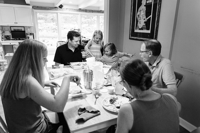 b/w extended family eats a meal at the dining room table together on summer nights