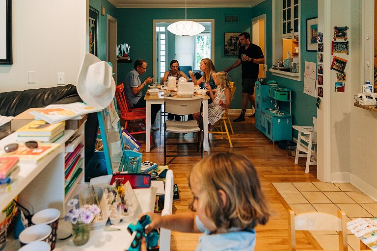 extended family members eat take out food in dining room while young son plays in adjacent area on summer nights