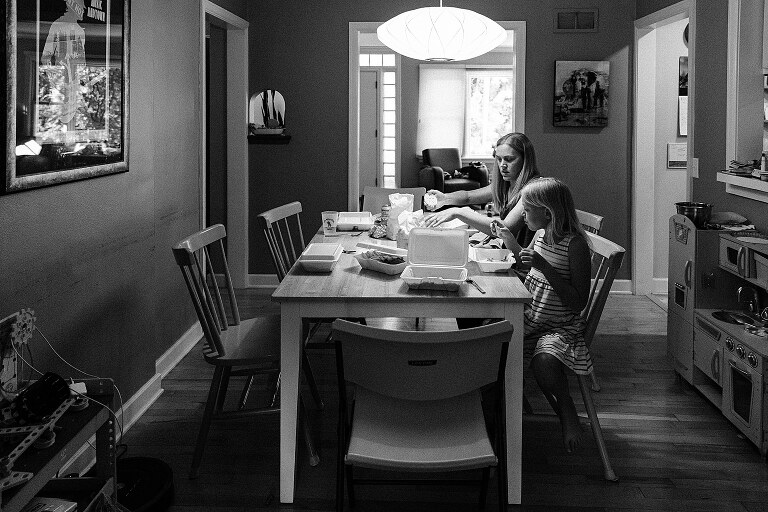 b/w mother and child sit under a light on dining room table opening take out food