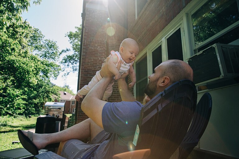 Dad holds baby up with sun shining as baby smiles at him while family packs house to be moving on