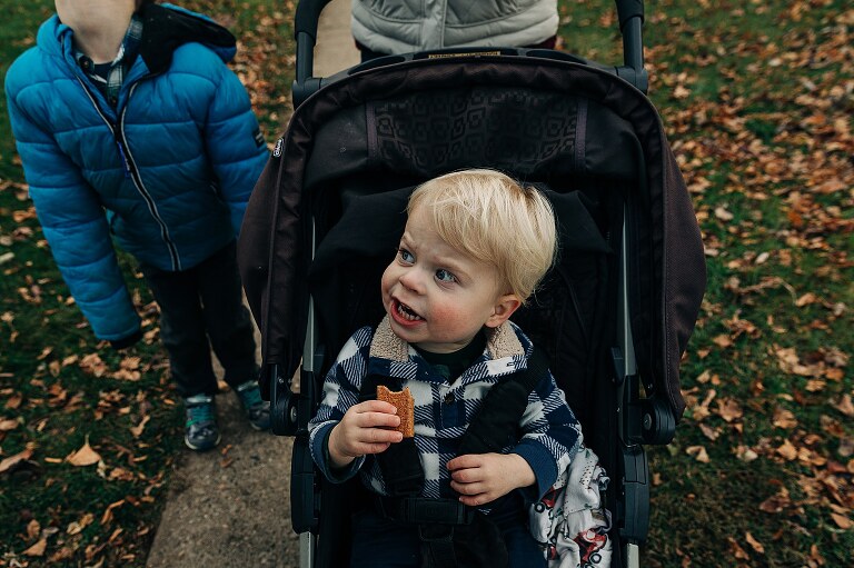 toddler in stroller and holding a snack makes a yelling face while older brother in blue coat is in the background together