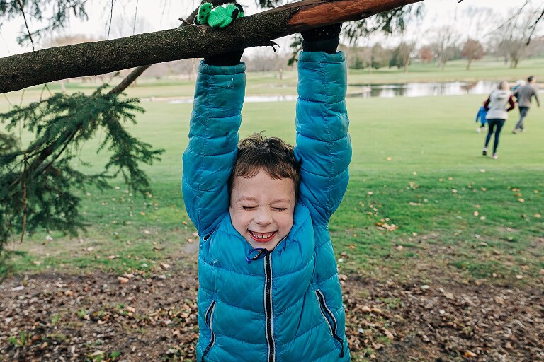 boy hangs from a brach and smiles with eyes closed while family runs toward water behind him