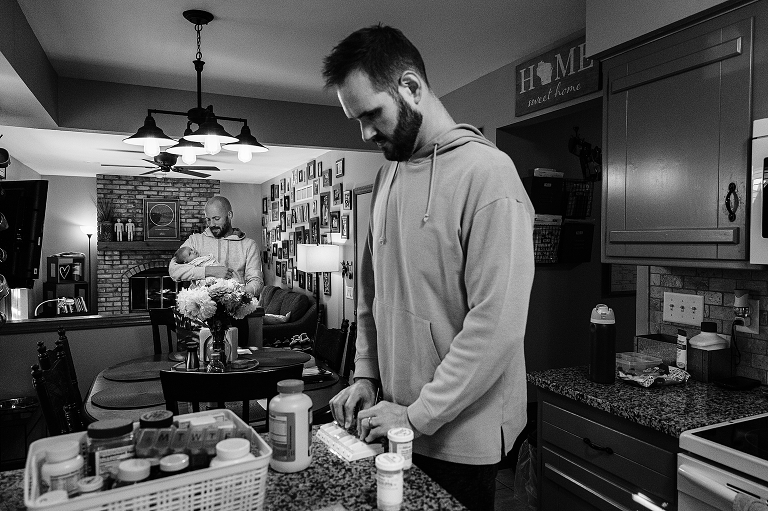 Black and white. father adds vitamins to weekly holder while other father holds newborn 