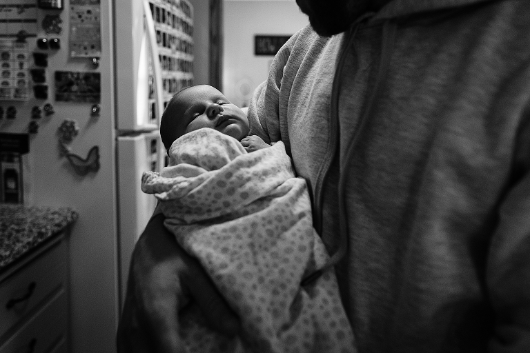 Black and white. father holds swaddled newborn in kitchen 