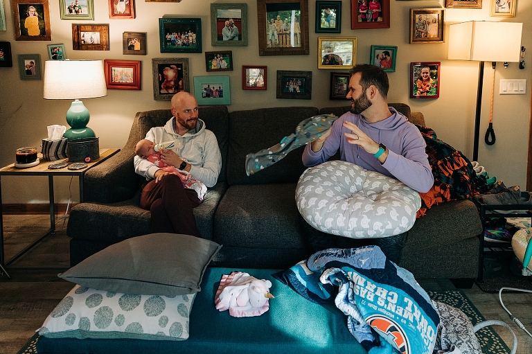 Fathers converse while feeding their newborn baby on the couch.