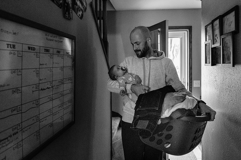 Black and white, father walks through hall with laundry basket and newborn baby