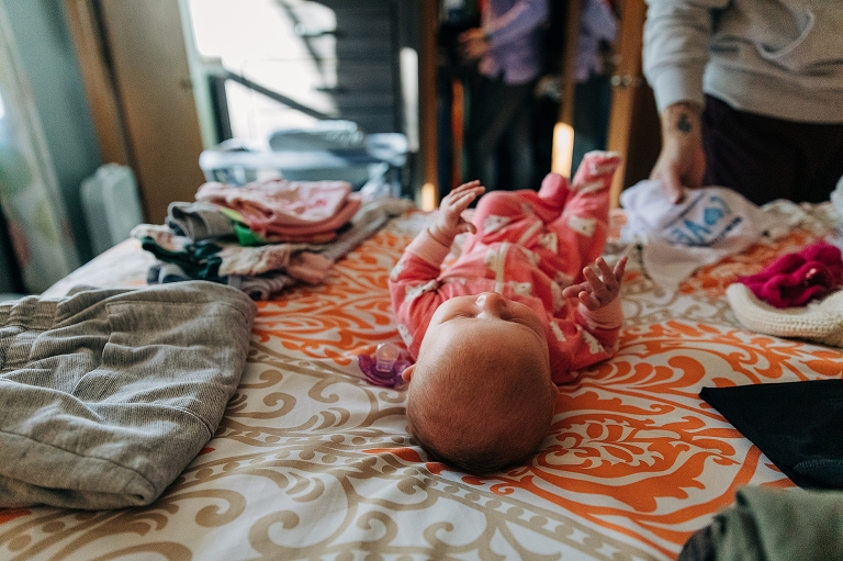 Newborn in pink onesie lays on bed next to laundry.