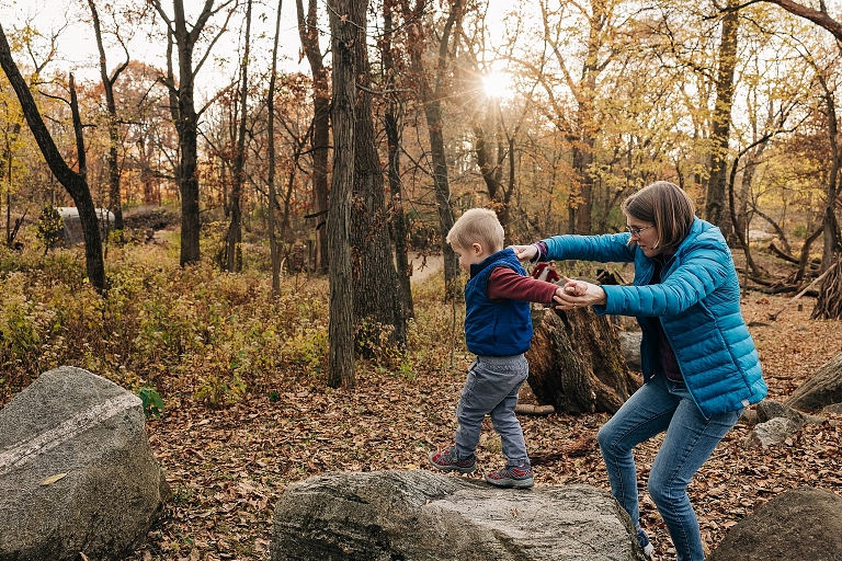 mothers help guide toddler son across big boulders while sun is shining through fall trees