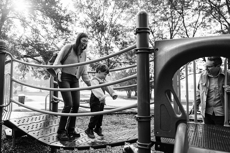 Mothers jump on a swingy bridge with her preschool age son at the park. Both are smiling. 