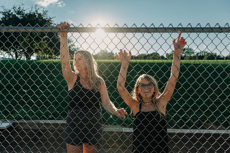 mothers and daughter stand at baseball diamond chainlink fence with sun setting behind them