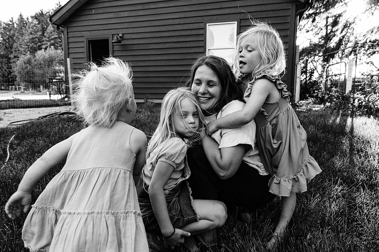 mothers sit in grass with three young daughters hugging and or running toward her