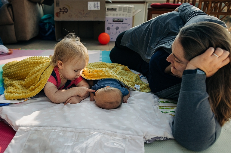 a mothers lay on her side watching her toddler daughter gently touch newborn son's nose.