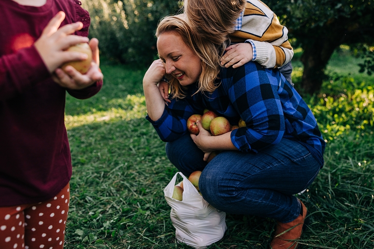 mothers squat holding an armful of apples while smiling and small child climbs on her back