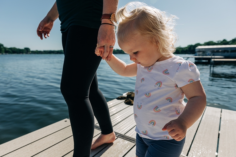 mom holds toddler daughter's hands while walking on the lake dock on a summer day. Toddler wears a rainbow shirt.