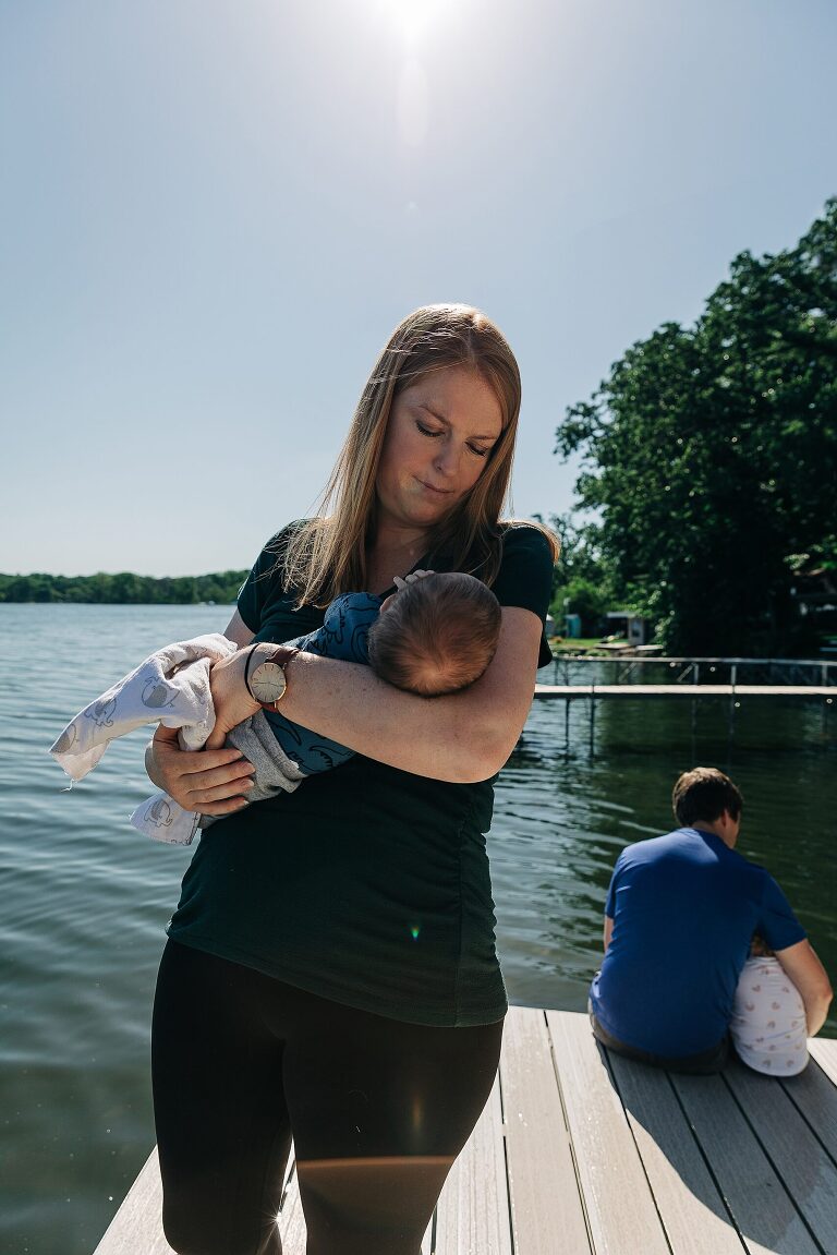 Mom cradles baby in her arms at the lake, while dad cradles toddler in the background.  Lake and sunshine 
