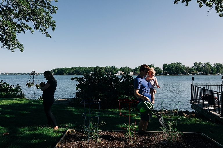 Dad and toddler water the tomato plants while mom and newborn walk in opposite direction. Lake is in the background. 