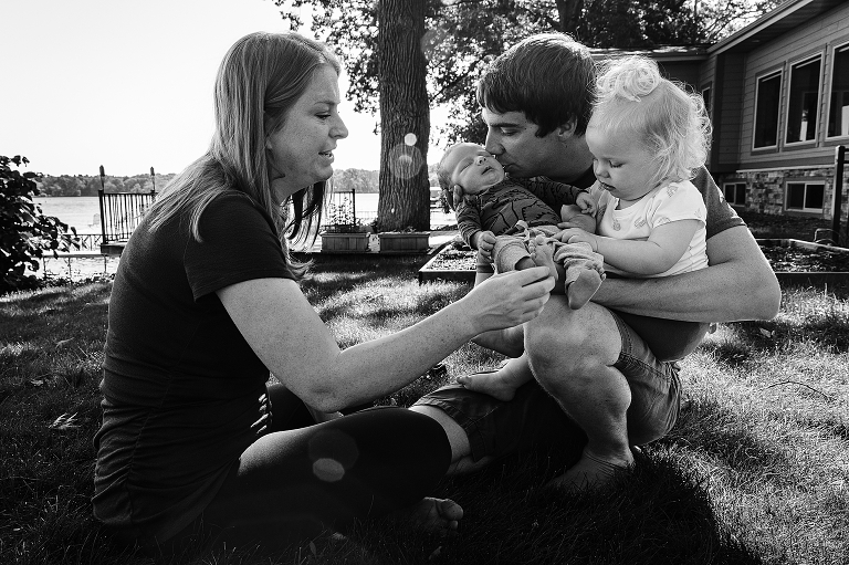 Family of four smile and connect with each other outside next to the lake. Dad kisses baby.