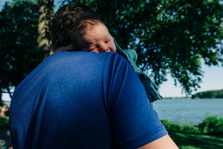 newborn on Dad's shoulder squints in the morning brightness of the day, lake is in the background 