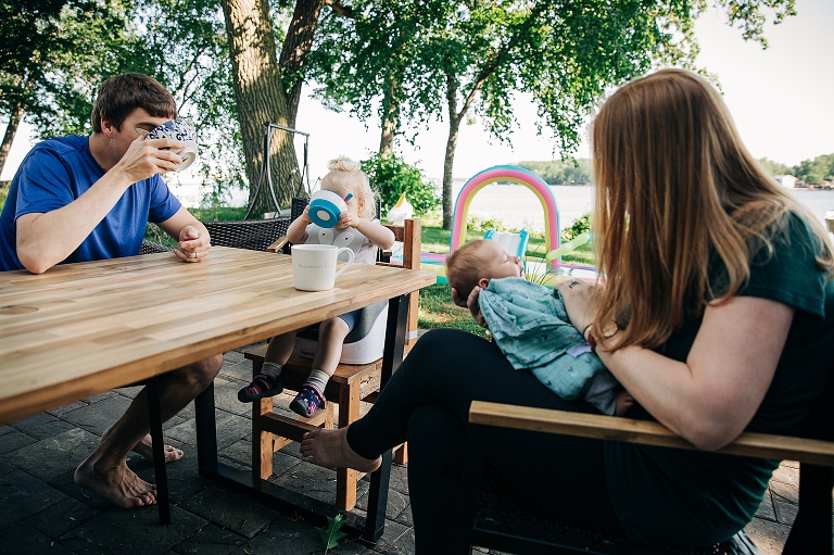 Dad and daughter both tip cereal bowls to cover their face and drink the milk while Mom stares lovingly at the newborn. Lake is in the background. 