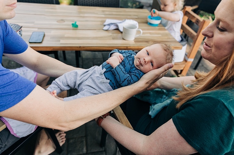 Dad passes newborn to mom while toddler girl eats cereal in the background 