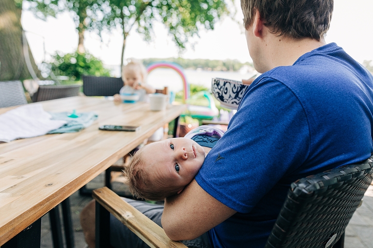 Focus on alert newborn face being held by dad eating cereal and toddler girl at the table