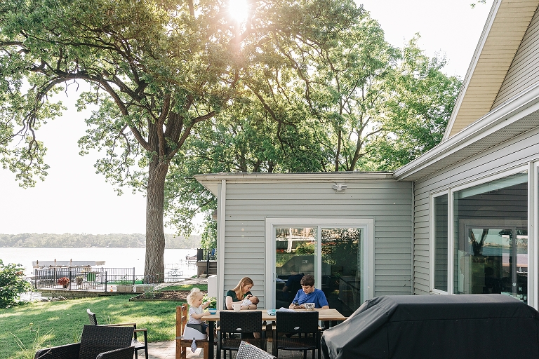 A family of four sits on an outside patio in the summer sun, eating breakfast together. The lake is nearby.