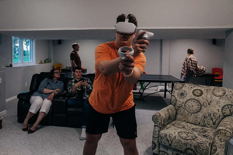 Youngest brother plays a game with a VR headset while mother and eldest look at edest's phone, and dad and daughter play ping pong in the background