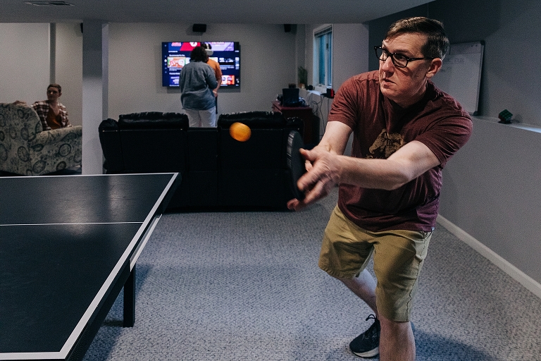 Father returns ping pong shot while adult daughter, mother and youngest brother browse the TV in the background, "all together now"