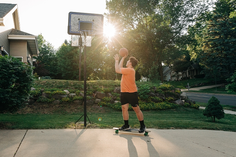 Youngest son attempts moving skateboard layup on outdoor basketball hoop with late afternoon sun in the background